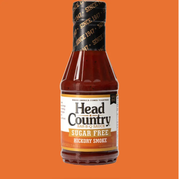 A close-up shot of a bottle of Head Country sugar-free hickory smoke BBQ sauce against an orange background.