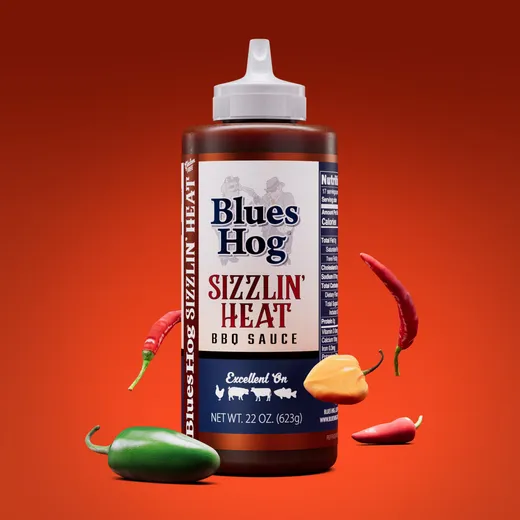 A clean shot of the Blues Hog Sizzlin' Heat BBQ Sauce bottle against a red background, with peppers floating around it.
